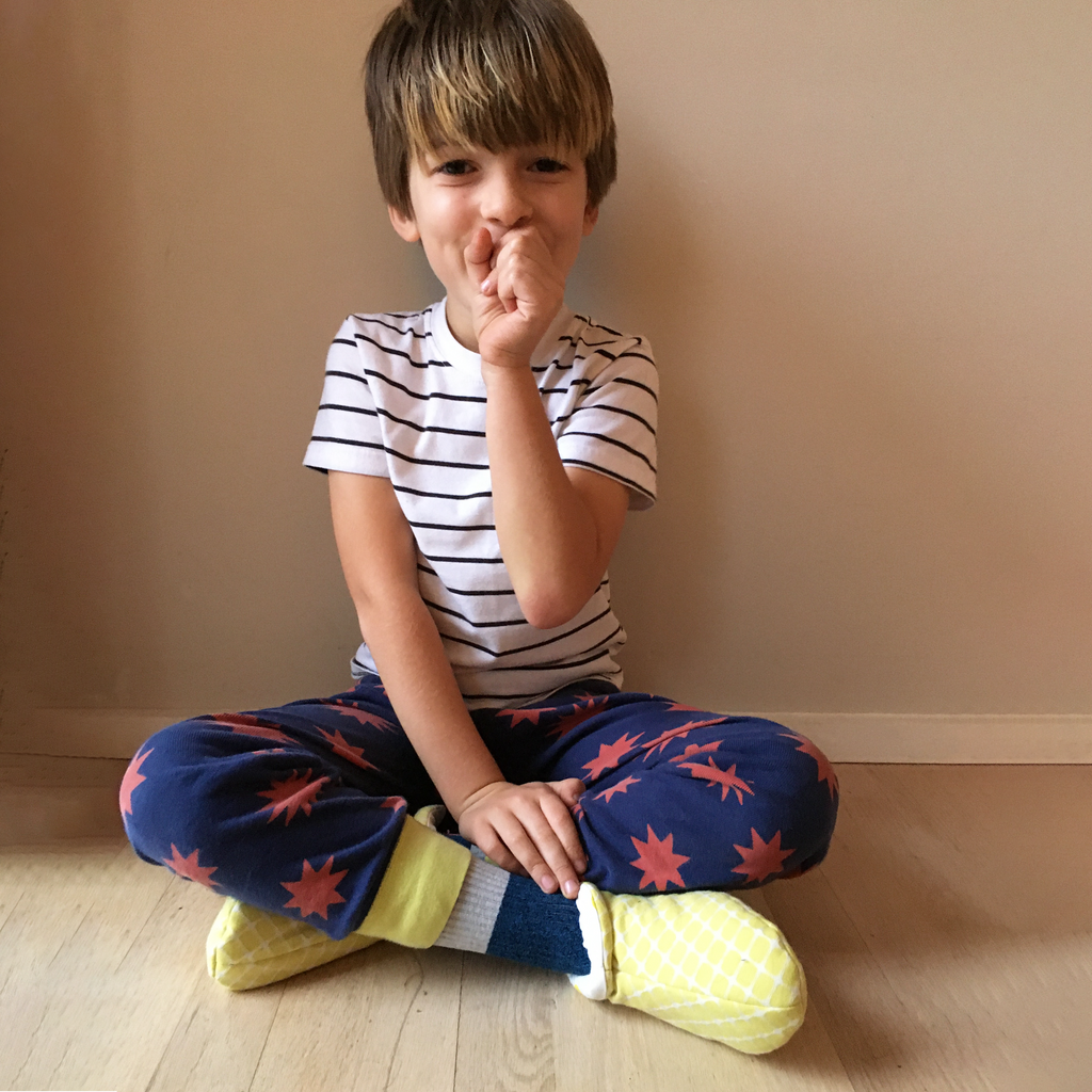 Kids Cozy Slipper sewing pattern — child sitting cross-legged wearing handmade slippers in yellow geometric print fabric with grey cuff, front view