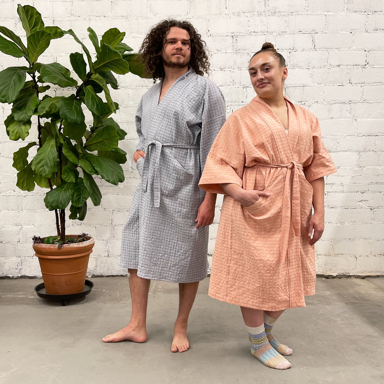 Slow Robe sewing pattern — two models wearing knee-length robes with kimono sleeves, tie waist, and patch pockets; blue-grey waffle fabric (left) and pink waffle fabric (right), side view