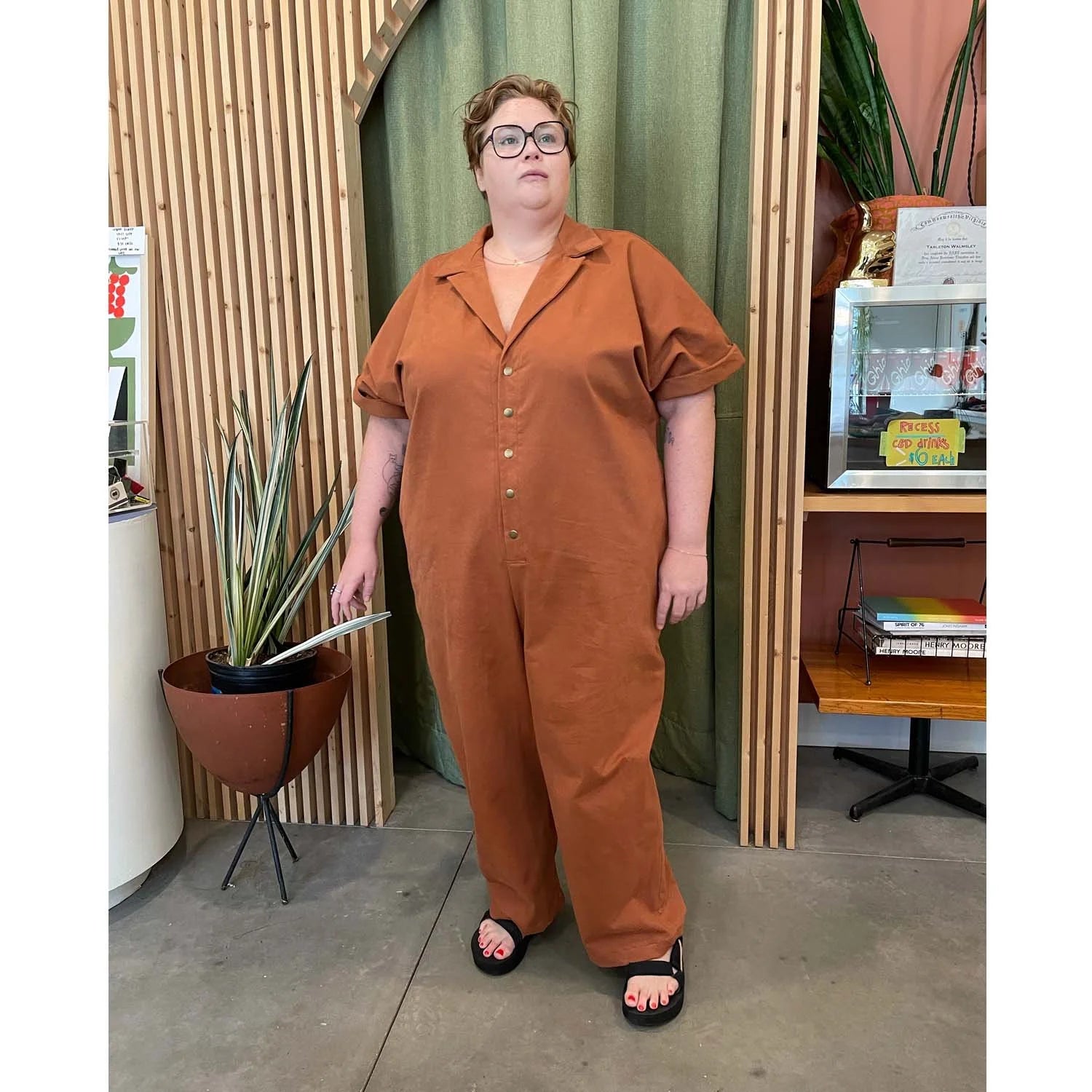 Bumper Bundle sewing pattern — Person wearing a rust colored jumpsuit standing in an indoor setting with plants and furniture - front relaxed view.