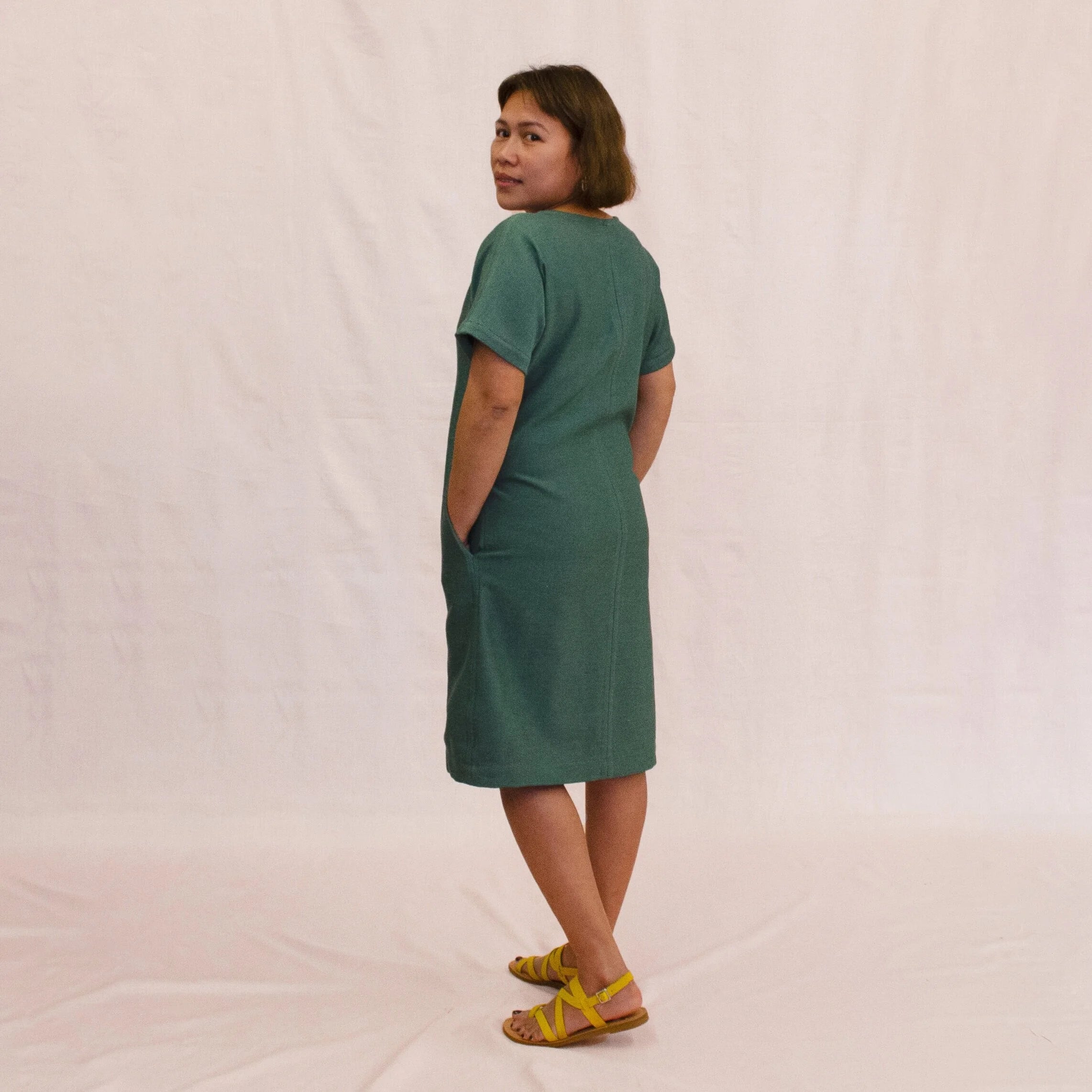 Day Dress sewing pattern — model wearing a relaxed knee-length shift dress in teal linen with scoop neckline, short sleeves, center front seam, and side pockets, back view