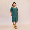 Day Dress sewing pattern — model wearing a relaxed knee-length shift dress in teal linen with scoop neckline, short sleeves, center front seam, and side pockets, front view