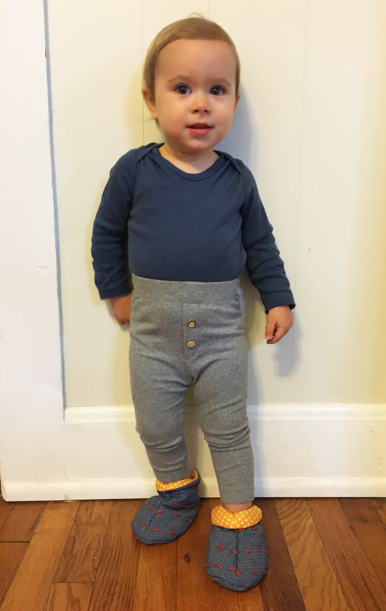 Kids Cozy Slipper sewing pattern — child sitting cross-legged wearing handmade slippers in navy geometric print fabric with grey cuff, front view